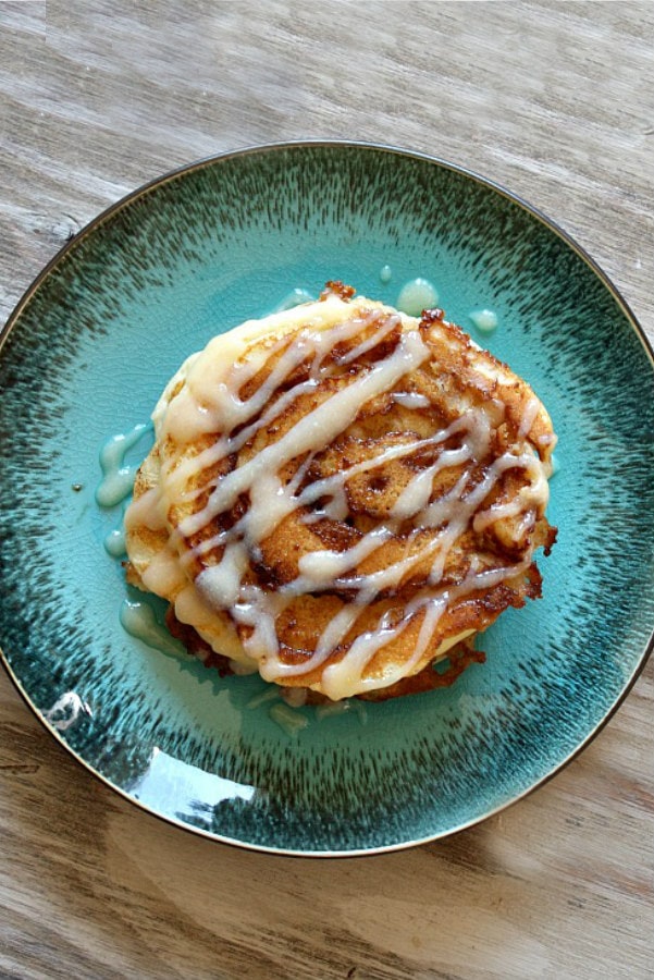 overhead shot of cinnamon roll pancakes with cream cheese drizzle on a teal green plate on a wooden board